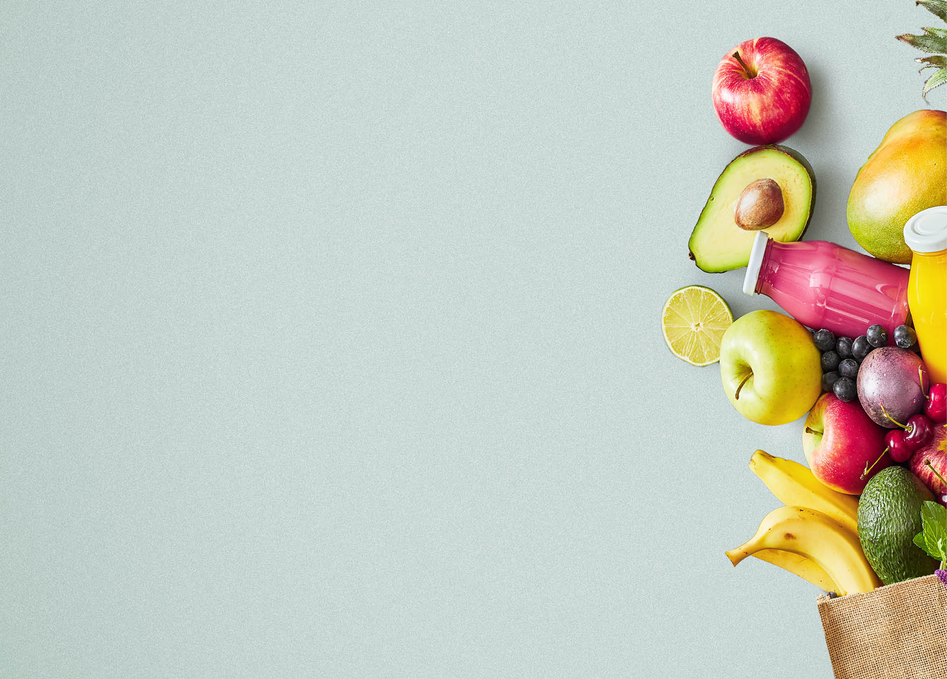 Reusable grocery bag of fresh healthy organic summer tropical fruit and bottles of juice spilling onto a green background in a flat lay still life panorama banner with copy space